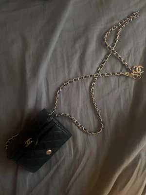 Svart plånbok med kedja - Snygg svart plånbok i quiltat läder med guldfärgade detaljer och ikonisk CC-logga. Plånboken har en lång kedja som gör att den kan bäras crossbody eller runt halsen. Perfekt accessoar för dig som gillar lyxig och tidlös stil.