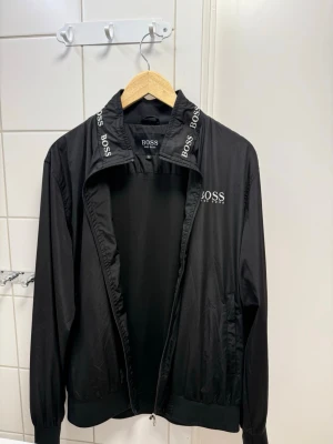 Svart vindjacka från BOSS - Stilren svart vindjacka från BOSS med vit logga på bröstet och tryckt BOSS-text på kragen. Jackan har dragkedja framtill, ribbade muddar och två sidofickor. Tillverkad i lätt och smidigt material som passar perfekt till streetwear.