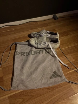 Adidas Predator elite vita fotbollsskor - Säljer ett par vita Adidas Predator elite fotbollsskor med grå detaljer och klassisk snörning. Skorna har dobbar för bra grepp på planen och kommer med en matchande Predator-påse. Perfekta för dig som vill ha stilrena och funktionella skor på matchen. Tyvärr har skorna lite defekter och skorna kommer inte med originell snörerna efter som att dom har gott sönder.