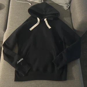Mörkblå hoodie från Russedress - Svart hoodie från Russedress med vit dragsko i huvan och stor magficka framtill. Klassisk passform och ribbade muddar vid ärmar och nederkant. Logga i vitt på ena ärmen. Perfekt för chill dagar och streetwear vibes. Passar folk med M bra