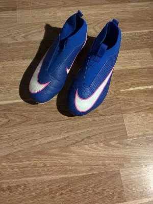 Blå Nike fotbollsskor utan snörning - Säljer ett par blå Nike fotbollsskor med stor vit och rosa swoosh på ovansidan. Skorna har en strömlinjeformad design utan snörning och dobbar i rosa och vitt för bra grepp på planen. Perfekta för dig som vill ha stilrena och moderna skor till matchen.