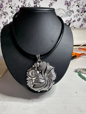 Stort halsband med silverfärgad blomma - Unikt halsband med en stor, rund silverfärgad hängberlock i form av en blomma med detaljerade mönster och små bollar. Hängs på ett svart, tjockt läderrem som ger en cool kontrast till det blanka silvret. Perfekt för dig som gillar statement-smycken och vill sticka ut.