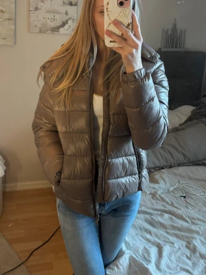 Brun pufferjacka med huva - Snygg  pufferjacka med huva och dragkedja framtill. Jackan har en quiltad design och är lätt vadderad för extra värme. Perfekt för kalla dagar och enkel att matcha med olika outfits. Passar dig som vill ha en stilren och bekväm vinterjacka. (Jackan ser något brun ut men den är mer lilabrun) 