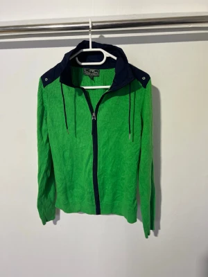 Grön vindjacka med marinblå detaljer lauren Ralph lauren - Grön vindjacka från lauren Ralph lauren med marinblå huva och axelparti. Jackan har dragkedja framtill, dragsko i huvan och långa ärmar. Materialet är lätt och krinklat, perfekt för blåsiga dagar. Snygg kontrast mellan färgerna ger en sportig look.