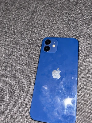 Blå iPhone 12 med dubbla kameror - Snygg blå iPhone 12 från Apple med stilren glasbaksida och två kameror på baksidan. Telefonen har en platt design med aluminiumram och Apples logga i silver. Perfekt för dig som gillar modern design och vill ha en kraftfull smartphone.OBS!! Priset kan diskuteras 