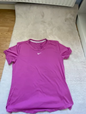 Rosa Nike t-shirt i funktionsmaterial - Snygg rosa t-shirt från Nike med klassisk logga på bröstet. Tillverkad i ett mjukt och lätt funktionsmaterial som andas, perfekt för träning. T-shirten har rund halsringning och korta ärmar för en sportig look.