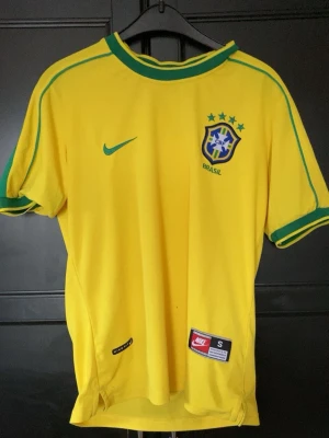 Brasilien Nike fotbollströja Kaká - Gul Brasilien fotbollströja från Nike med gröna detaljer, officiellt klubbmärke och fyra stjärnor på bröstet. Tryck med Kaká och nummer 8 på ryggen. Tillverkad i lätt och ventilerande material, perfekt för match eller träning.