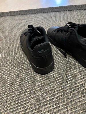 Svarta Adidas sneakers i läder - Snygga helsvarta sneakers från Adidas med klassisk låg profil. Skorna har ovandel i slätt läder, svarta skosnören och diskret Adidas-logga på hälen. Perforerade detaljer på sidan för extra stil och komfort.