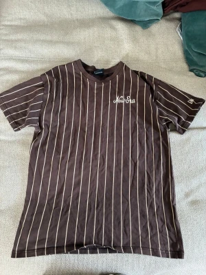 Randig brun t-shirt från New Era - Snygg brun t-shirt från New Era med vita vertikala ränder och broderad logga på bröstet. Lite oversized passform och rund halsringning. Perfekt för en avslappnad streetwear-look. Minimal lagning, se bild. Är storlek XS men är som sagt oversized så passar som S. Priset kan diskuteras.