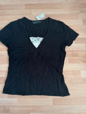 Ny. Svart omlottopp med spetsdetalj - Svart omlottopp med korta ärmar och v-ringning. Toppen har en vit spetsdetalj vid halsen som ger en snygg kontrast. Perfekt för dig som gillar en enkel men ändå stylish look.