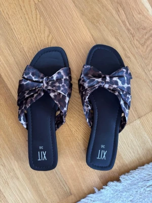 Leopardmönstrade sandaler med rosett - Säljer ett par trendiga sandaler från XIT i storlek 36. De har leopardmönstrad textil med stora rosetter framtill och svart sula. Perfekta för dig som vill sticka ut med ett djurmönster och gillar en bekväm slip-in modell.