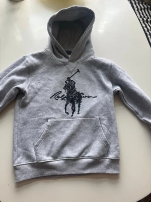 Grå hoodie från Ralph Lauren - Snygg grå hoodie från Ralph Lauren med stor svart polospelare och logga på bröstet. Klassisk känguruficka framtill och huva. Tillverkad i mjukt bomullsmaterial som är perfekt för en avslappnad och trendig look.