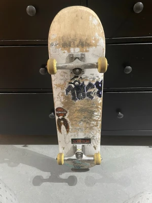 Skateboard med vita hjul och stickers - Säljer en skateboard med vit undersida dekorerad med flera stickers, bland annat en fjäder och text. Brädan har gula hjul och silverfärgade trucks. Ovansidan har svart griptape. Perfekt för dig som gillar street och skate.jag kommer inte att skicka,för det är svårt att hitta lång låda . Jag kan bara möta er i Stockholm 