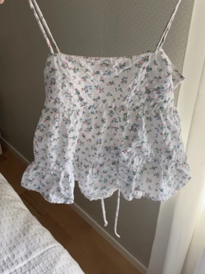 Blommig vit linnetopp med volang - Supersöt vit linnetopp med små blommor i pastellfärger. Toppen har tunna axelband, volangkant nertill och snörning i ryggen för en justerbar passform. 