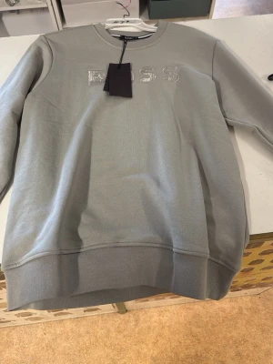 Ljusgrå sweatshirt från BOSS - Snygg ljusgrå sweatshirt från BOSS med broderad logga på bröstet. Tröjan har rund halsringning och ribbade muddar vid ärmslut och nederkant. Tillverkad i mjukt bomullsmaterial som känns skönt mot huden. Perfekt för en clean och stilren look.