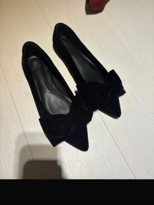 Svarta ballerinaskor med rosett - Stilrena svarta ballerinaskor från Next med spetsig tå och stor sammetsrosett framtill. Storlek 41, passar mig som vanligtvis har 40. Oanvända!
