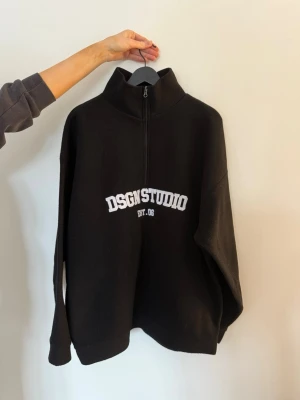 Svart halfzip hoodie DSGN STUDIO - Svart tröja med vit DSGN STUDIO broderad text på bröstet och halv dragkedja framtill. Oversized passform och hög krage ger en chill vibe. Perfekt till jeans eller mjukisbyxor. Materialet känns mjukt och bekvämt mot huden.