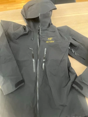 Svart vindjacka från Arc'teryx Large  - Snygg svart vindjacka från Arc'teryx med huva och dragkedja framtill. Jackan har flera vattentäta fickor med dragkedja, justerbara muddar med kardborre och gul Arc'teryx-logga på bröstet. Perfekt för blåsiga dagar och outdoor-äventyr.