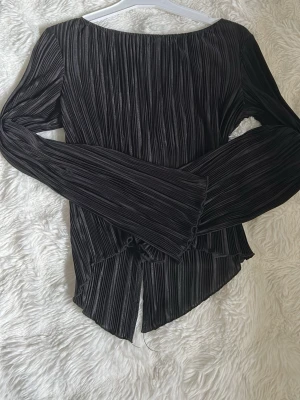 Svart plisserad topp med knyt - Trendig svart plisserad topp med långa ärmar och snygga knyt baktill. Toppen har en lätt glansig finish och asymmetrisk nederkant som ger en cool look. Perfekt att styla med jeans eller kjol för en modern vibe.