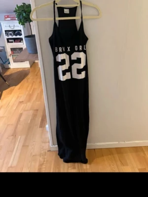 Svart långklänning Gina Tricot - Sportig svart långklänning från Gina Tricot med tunna axelband och tryck i vitt framtill med texten 'BRX GRL' och siffran 22. Klänningen har en rak passform och är tillverkad i ett mjukt, tunt material som känns skönt mot huden.