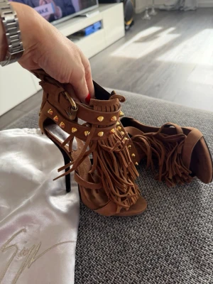 Bruna pumps med fransar och nitar - Säljer ett par bruna pumps från Giuseppe Zanotti i mocka med höga smala klackar. Skorna har fransdetaljer framtill och guldfärgade nitar längs remmarna. Öppen tå och spänne vid ankeln ger en cool och trendig look. Storlek 37.5