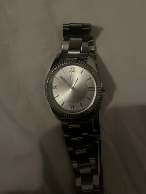 Klassisk silverfärgad armbandsklocka - Stilren armbandsklocka i helsilver med metallarmband och urtavla med romerska siffror. Klockan har en rund form och minimalistisk design utan logotyp eller extra funktioner. Perfekt accessoar för en clean look.