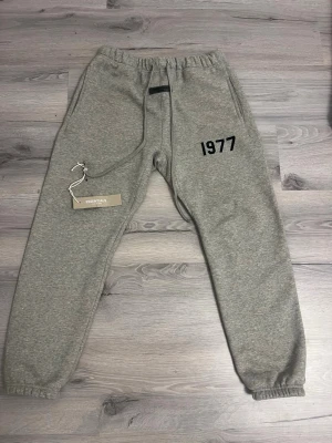 Grå mjukisbyxor Essentials 1977 - Snygga grå mjukisbyxor från Essentials Fear of God med trycket '1977' på vänster lår. Byxorna har resår i midjan och bensluten samt dragsko för bästa passform. Tillverkade i mjukt och bekvämt material, perfekta för chill dagar.