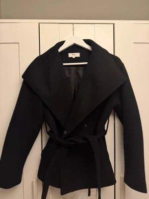 Svart kappa från Gina Tricot XS - Stilren svart kappa från Gina Tricot i ullblandning. Väldigt bra skick inga tecken på användning.  Det  Storlek Xs och köptes för 700kr.  Kappan har bred krage, knytband i midjan och en klassisk, rak passform.