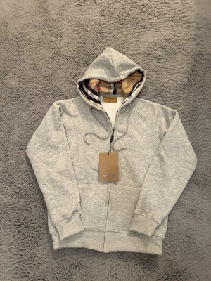 Grå hoodie från Burberry med dragkedja - Säljer en grå hoodie från Burberry med klassiskt rutigt foder i huvan. Hoodien har dragkedja framtill, snörning i huvan och ribbade muddar vid ärmar och nederkant. Perfekt för en chill och stilren look. Tröjan har bara blivit testad, aldrig använt. (Storlek M men passar bättre S. 160-175cm)