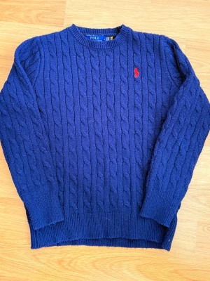 Mörkblå stickad tröja Polo Ralph Lauren - Snygg mörkblå stickad tröja från Polo Ralph Lauren med klassiskt kabelstickat mönster och röd broderad logga på bröstet. Rund hals och långa ärmar, perfekt för dig som gillar stilrena och tidlösa plagg. Passar dig som vill ha en chill men ändå preppy look.