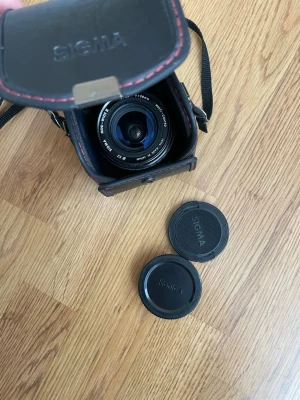 SIGMA vidvinkelobjektiv med väska - Säljer ett SIGMA vidvinkelobjektiv 24mm f/2.8 med Kenko och SIGMA objektivlock samt en svart läderväska med röd söm. Objektivet är kompakt, svart och har tydliga markeringar för bländare och fokus. Perfekt tillbehör för din kamera.