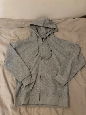 Grå zip hoodie från Polo Ralph Lauren - Säljer en grå hoodie med dragkedja från Polo Ralph Lauren. Klassisk design med luva och dragkedja, ribbade muddar och fickor framtill. Ikoniska Polo-loggan broderad på bröstet. Perfekt för chill dagar och enkel att matcha med allt.