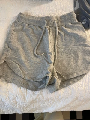 Grå mjukisshorts med snörning - Grå mjukisshorts i bomull med bred resår i midjan och snörning framtill. Shortsen har en avslappnad passform och rundade benslut för en sportig look. Perfekta för lata dagar eller träning.