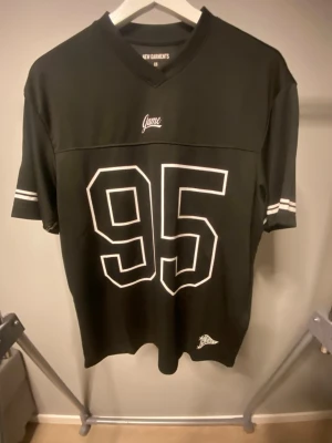 Svart t-shirt med tryck från New Garments - Snygg svart t-shirt från New Garments med stort vitt 95-tryck på framsidan och vita ränder på ärmarna. T-shirten har v-ringad hals och sportig look, perfekt för dig som gillar streetwear. Materialet känns som mjuk bomull eller polyester.