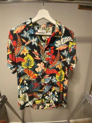 Blommig kortärmad skjorta - Färgstark blommig skjorta från bershka med svart bas och mönster i rött, gult, blått och vitt. Skjortan har korta ärmar, klassisk krage och knappar framtill. Perfekt för dig som vill sticka ut med en tropisk vibe.