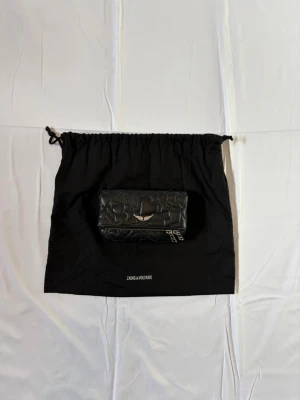 Svart väska från Zadig & Voltaire - Cool svart väska från Zadig & Voltaire i quiltat läder med präglade hjärtan, stjärnor och logga. Väskan har dragkedja och detaljer i silver. Perfekt för dig som gillar edgy accessoarer med unik design.