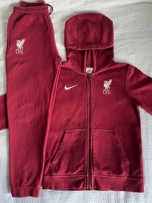 Liverpool FC/Nike träningsset barn M (137-147 cm) – hoodie + byxa - Snyggt träningsset från Nike / Liverpool FC i rött. Hoodie med dragkedja + matchande byxa.  Storlek: M (137–147 cm). Fint använt skick, inga synliga defekter.  Mjukt och skönt material – passar för träning eller vardag. Loggor fram på både tröja och byxa.  Säljs som set.