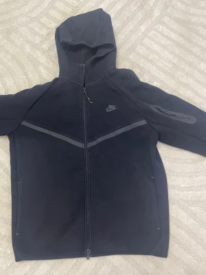 Nike tech  - Snygg svart hoodie från Nike med hel dragkedja framtill och huva. Hoodien har diskreta grå detaljer över bröstet och på ena ärmen samt Nike-logga på bröstet. Tillverkad i mjukt material som passar perfekt till en avslappnad streetwear-look.