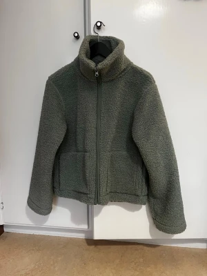 Grön teddyjacka från 157 - Mysig grön teddyjacka från 157 med hög krage och dragkedja framtill. Jackan har två stora fickor och är gjord i fluffigt teddy-material som håller dig varm. Perfekt för kyliga dagar och ger en avslappnad, trendig look. Varan är som ny, använd ett få tal gånger!!💕