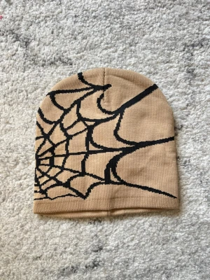 Beige mössa med spindelnätsmönster - Säljer en beige mössa med ett stort svart spindelnätsmönster. Mössan är i mjukt, stretchigt material och har en klassisk, rundad form. Perfekt för dig som vill sticka ut med en unik accessoar.
