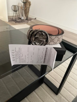 Gucci beige GG Supreme bälte - Säljer ett klassiskt Gucci GG Supreme bälte i beige med det ikoniska GG-mönstret och stor metallspänne framtill. Bältet har en lyxig känsla med detaljer i brunt och insida i ljusrosa. Perfekt accessoar för att lyfta vilken outfit som helst.