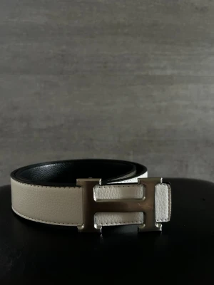 Hermess bälte - Stilrent beige skärp med ikoniskt H-format metallspänne. Bältet har en slät, elegant yta i skinn och svarta detaljer på insidan. Perfekt accessoar för att lyfta vilken outfit som helst.