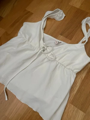 Vit linnetopp med knytband - Söt vit linnetopp med tunna axelband och knytband framtill. Toppen har en lätt rynkad midja och luftig passform, perfekt för varma dagar. Materialet känns mjukt och svalt mot huden.