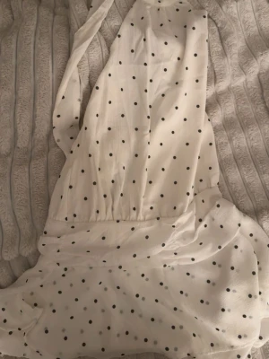 Vit prickig omlottklänning - Söt vit kortklänning med svarta prickar och omlottmodell. Klänningen har volangdetaljer och knytband i midjan. Tillverkad i ett lätt och luftigt material, perfekt för varma dagar. Djup v-ringning framtill och kort kjol med volangkant. Prislapp kvar.