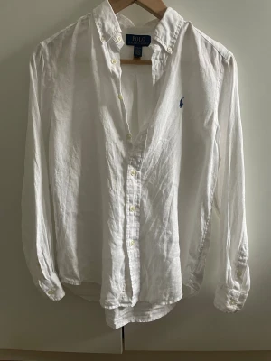 Vit linneskjorta från Polo Ralph Lauren - Klassisk vit skjorta från Polo Ralph Lauren i luftigt linne. Skjortan har lång ärm, knappar framtill och den ikoniska blå loggan broderad på bröstet. Perfekt för en clean och stilren look. Storlek 14-16 år motsvarar XS ungefär