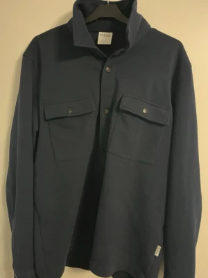 Mörkblå overshirt från Selected Homme - Stilren mörkblå overshirt från Selected Homme med två stora bröstfickor med lock och tryckknappar. Overshirten har klassisk krage, raka ärmar och är tillverkad i ett mjukt tyg som ger en avslappnad look. Perfekt att bära öppen eller stängd.