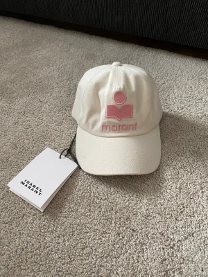 Vit keps från Isabel Marant - Snygg vit keps från Isabel Marant med broderad rosa logga och text framtill. Klassisk böjd skärm och justerbar baksida. Tillverkad i ett tåligt bomullstyg som ger en clean och fräsch look. Perfekt accessoar för att lyfta din outfit.