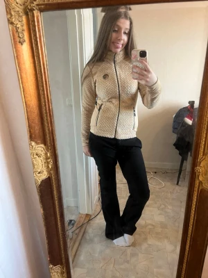 Beige teddytröja från J. Lindeberg - Säljer en beige teddytröja från J. Lindeberg med dragkedja framtill och två fickor med dragkedja. Tröjan har en mjuk, fluffig framsida och släta ärmar. Märkets logga i metall sitter på bröstet. Perfekt för dig som gillar sportig och trendig stil.