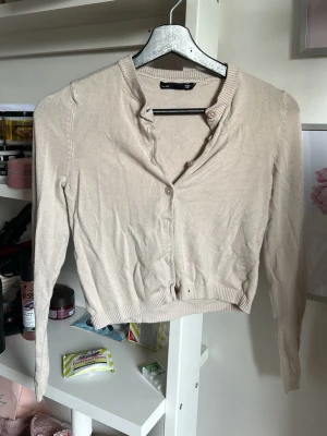 Beige cardigan från 157 - Snygg beige cardigan från 157 med långa ärmar och ribbade muddar. Modellen har v-ringning och knäppning framtill med små knappar. Perfekt att slänga över en topp för en enkel och clean look.