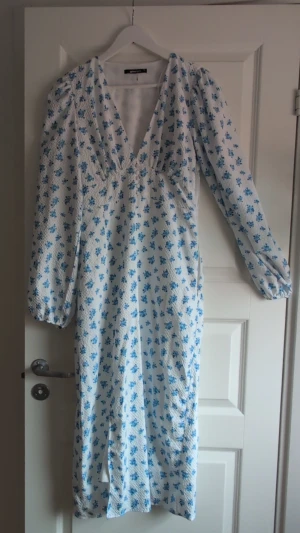 Vit blommig långklänning Gina Tricot - Långklänning från Gina Tricot i vitt med blått blommönster. Klänningen har v-ringning, långa puffärmar och en figurnära passform upptill. Perfekt för sommarens alla tillfällen.